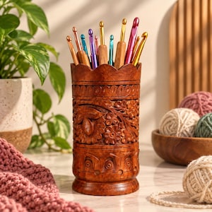 May include: A cylindrical wooden holder with a scalloped top edge and intricate floral carvings, filled with a variety of colorful metal and wooden crochet hooks in shades of pink, blue, gold, and brown, standing upright on a flat surface.