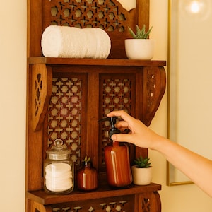 Puede incluir: Estanter&iacute;a de ba&ntilde;o de madera con intrincados dise&ntilde;os calados. Contiene una toalla blanca enrollada, una peque&ntilde;a planta en maceta, un tarro de cristal con discos de algod&oacute;n y una botella marr&oacute;n con bomba negra. La estanter&iacute;a est&aacute; montada en una pared clara.