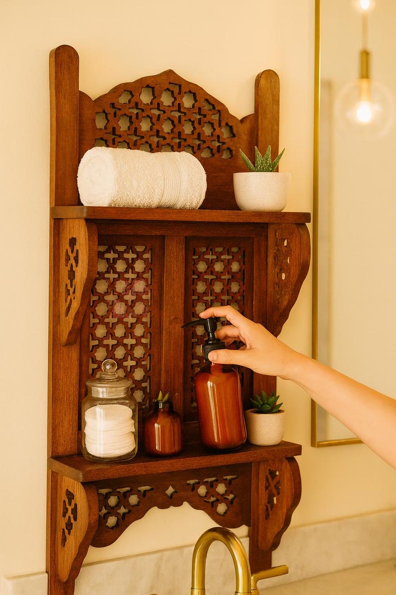 Puede incluir: Estanter&iacute;a de ba&ntilde;o de madera con intrincados dise&ntilde;os calados. Contiene una toalla blanca enrollada, una peque&ntilde;a planta en maceta, un tarro de cristal con discos de algod&oacute;n y una botella marr&oacute;n con bomba negra. La estanter&iacute;a est&aacute; montada en una pared clara.
