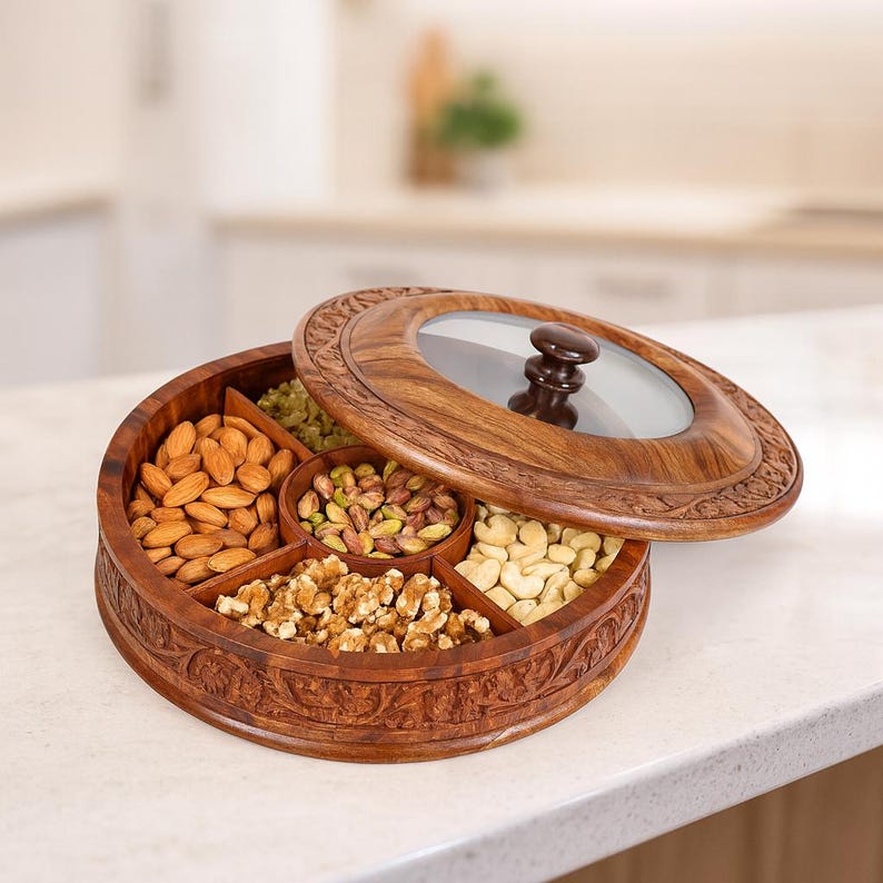 May include: A circular wooden serving tray with a glass lid. The tray is divided into five compartments, each containing different nuts. The tray is made of dark brown wood and has carved floral patterns around the rim.