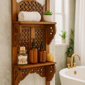 Puede incluir: Estanter&iacute;a de ba&ntilde;o de madera con detalles tallados. La estanter&iacute;a contiene una toalla blanca enrollada, una peque&ntilde;a planta en maceta, dos botellas con bomba marrones, un tarro de bastoncillos de algod&oacute;n y una esponja natural. La estanter&iacute;a est&aacute; montada en una pared de baldosas de m&aacute;rmol.