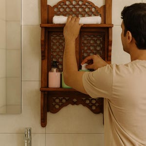 Puede incluir: Un estante de ba&ntilde;o de madera oscura con intrincados dise&ntilde;os calados. El estante contiene una toalla blanca enrollada y dos botellas. Una persona est&aacute; alcanzando un art&iacute;culo en el estante.