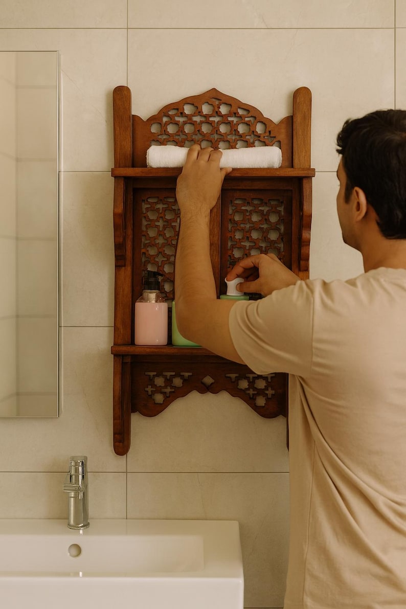 Puede incluir: Un estante de ba&ntilde;o de madera oscura con intrincados dise&ntilde;os calados. El estante contiene una toalla blanca enrollada y dos botellas. Una persona est&aacute; alcanzando un art&iacute;culo en el estante.