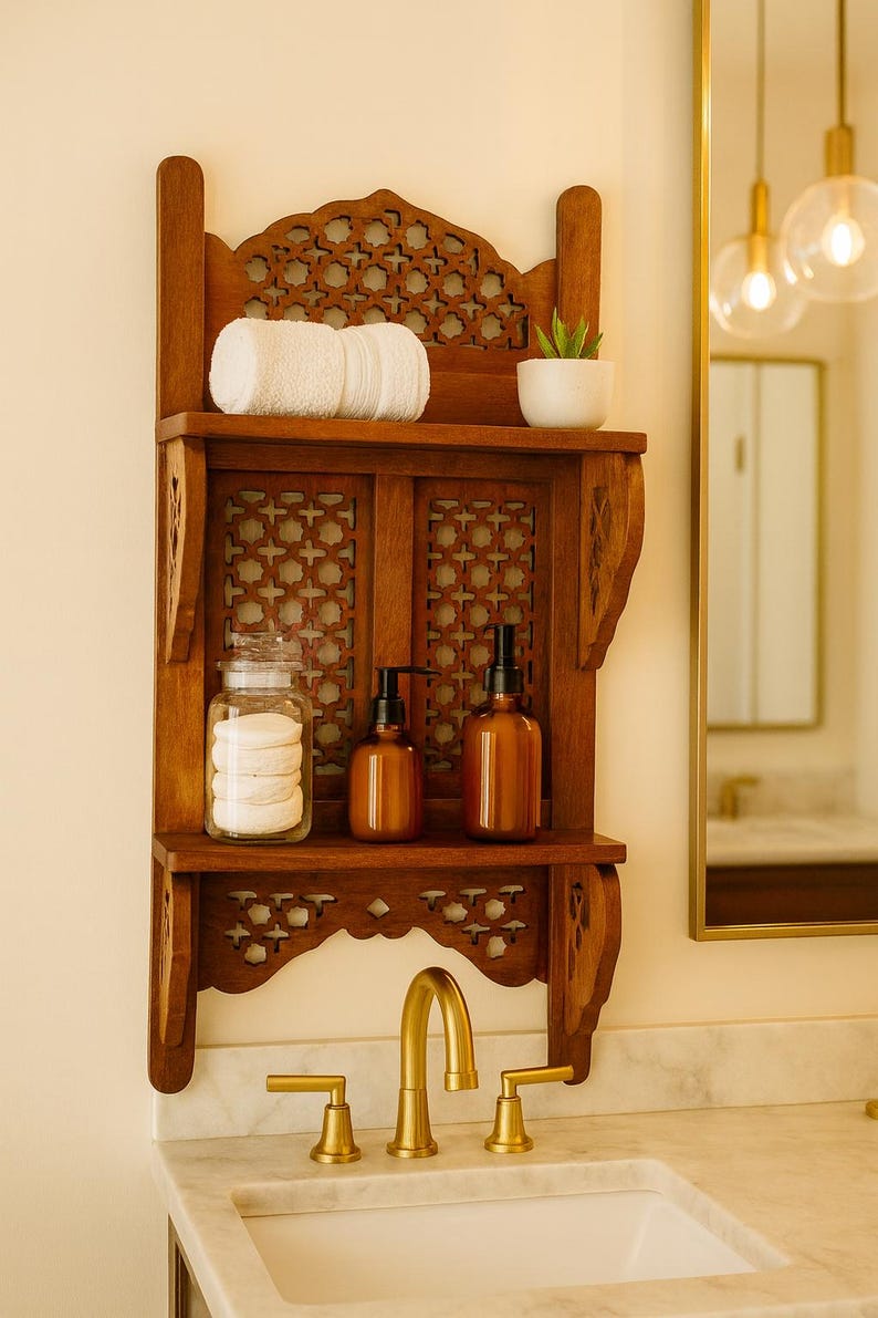 Puede incluir: Estanter&iacute;a de ba&ntilde;o de madera con intrincados dise&ntilde;os calados. Contiene una toalla blanca enrollada, una peque&ntilde;a planta en maceta, un tarro de discos de algod&oacute;n y dos botellas &aacute;mbar con dispensador. Un grifo dorado en primer plano.