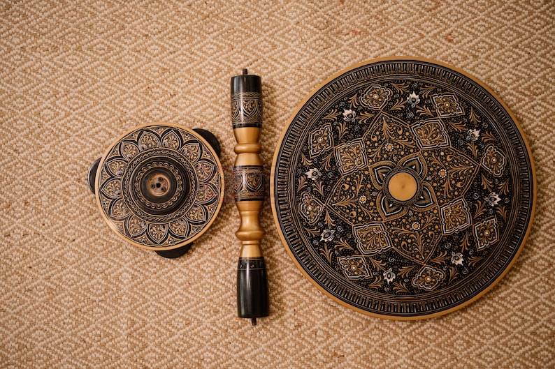 Pu&ograve; includere: Un set di tre elementi decorativi in legno con intricati motivi floreali neri e dorati. Il set comprende un piano del tavolo rotondo, un pezzo circolare pi&ugrave; piccolo e una colonna di supporto centrale. I pezzi sono esposti su una superficie strutturata marrone chiaro.