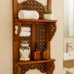 Puede incluir: Estanter&iacute;a de ba&ntilde;o de madera con intrincados detalles tallados. Contiene toallas blancas enrolladas, una peque&ntilde;a planta en maceta y frascos de vidrio con bastoncillos de algod&oacute;n. Tambi&eacute;n hay dos botellas con dosificador &aacute;mbar.