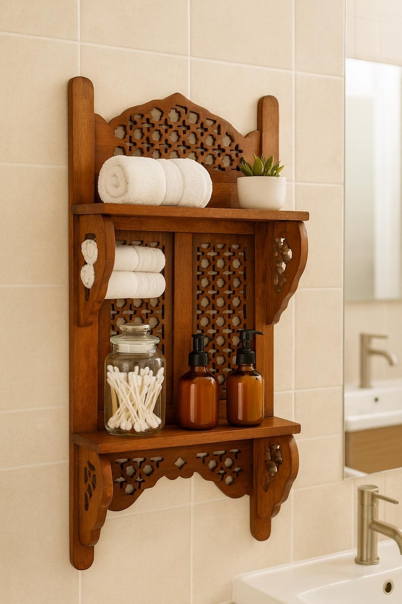 Puede incluir: Estanter&iacute;a de ba&ntilde;o de madera con intrincados detalles tallados. Contiene toallas blancas enrolladas, una peque&ntilde;a planta en maceta y frascos de vidrio con bastoncillos de algod&oacute;n. Tambi&eacute;n hay dos botellas con dosificador &aacute;mbar.