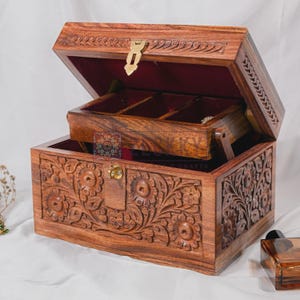 May include: A wooden jewelry box with intricate floral carvings. The box is open, revealing a red velvet interior and a smaller, tiered compartment. It features a brass clasp and hinges. The wood has a warm, reddish-brown tone.