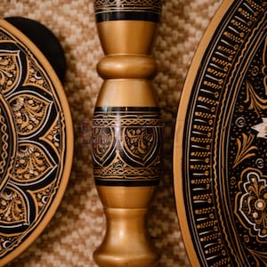 Pu&ograve; includere: Primo piano di oggetti decorativi in legno con intricati disegni neri e dorati. L'immagine presenta un pilastro centrale ornato e due piatti circolari con motivi dettagliati, che mostrano una miscela di artigianato e arte.