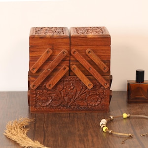 May include: A wooden sewing box with a carved floral design. The box is a warm brown color and has multiple compartments that open and close. A perfume bottle and a necklace are also visible.