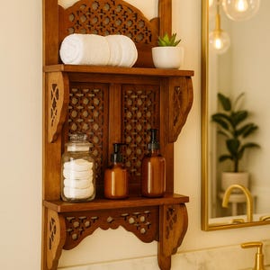 Puede incluir: Estanter&iacute;a de ba&ntilde;o de madera con intrincados detalles tallados. La estanter&iacute;a contiene una toalla blanca enrollada, una peque&ntilde;a planta en maceta, un tarro de cristal con discos de algod&oacute;n y dos botellas con dosificador &aacute;mbar. La estanter&iacute;a est&aacute; montada en una pared clara.