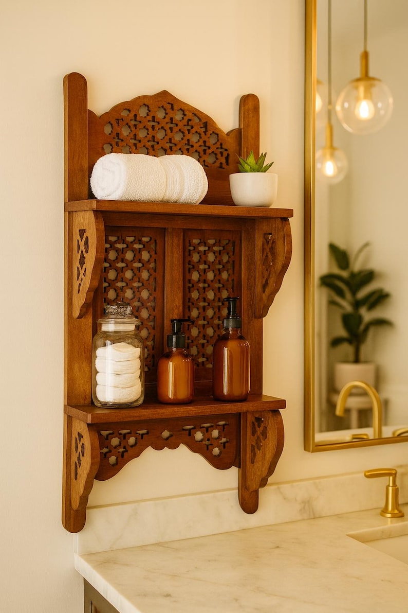 Puede incluir: Estanter&iacute;a de ba&ntilde;o de madera con intrincados detalles tallados. La estanter&iacute;a contiene una toalla blanca enrollada, una peque&ntilde;a planta en maceta, un tarro de cristal con discos de algod&oacute;n y dos botellas con dosificador &aacute;mbar. La estanter&iacute;a est&aacute; montada en una pared clara.