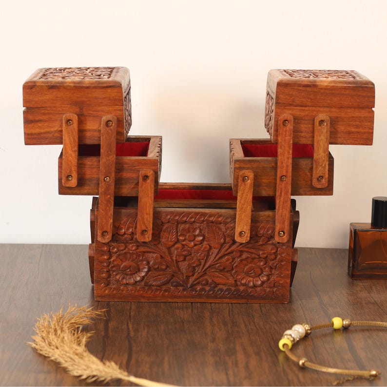 May include: A handcrafted wooden jewelry box with a unique, tiered design. The box features intricate floral carvings and a rich brown finish. The interior is lined with red fabric. The box has two hinged compartments that open upwards.