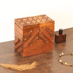 May include: A wooden, multi-tiered box with a hinged lid and intricate diamond-shaped carvings. The box is a warm brown color and has several compartments. A small brown bottle and a beaded necklace are also visible.