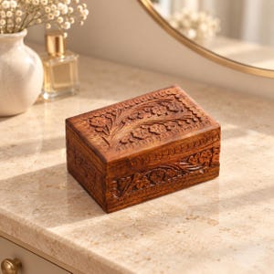 May include: A rectangular wooden box with a dark brown finish, featuring intricate hand-carved floral patterns on the lid and front panel. The box has a hinged lid and a smooth, polished surface that highlights the natural wood grain texture.