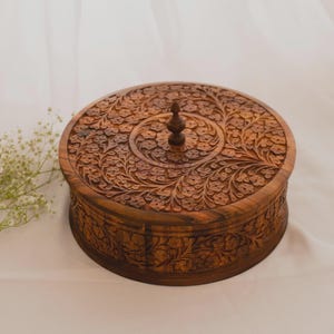 May include: A round, wooden box with a lid, intricately carved with floral patterns. The box is a warm brown color and features a small, decorative knob on the lid. The design is symmetrical and detailed, suggesting a handcrafted item.