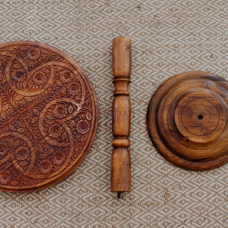 May include: A disassembled wooden table with a carved floral design on the tabletop. The table has a central support column and a base. The wood has a warm, brown tone. The pieces are arranged on a textured, beige surface.