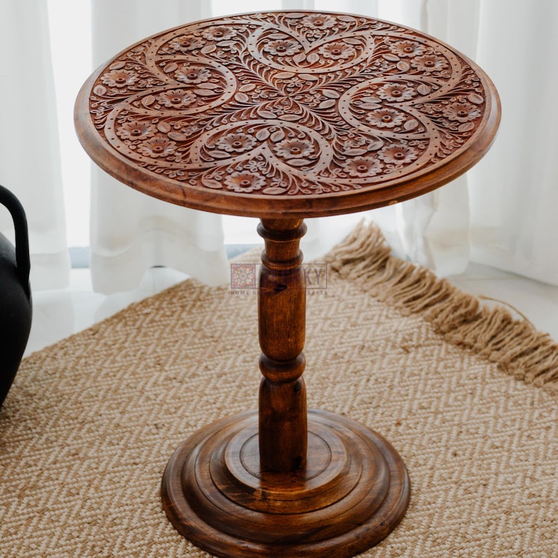May include: A round, dark brown wooden side table with intricate floral carvings on the tabletop. The table has a single, turned pedestal leg and a circular base. The table is approximately 24 inches tall.