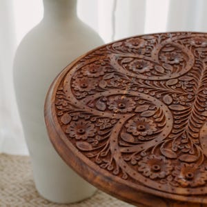 May include: A round, wooden table with intricate floral carvings. The table is a warm brown color and features detailed patterns of flowers and leaves. A white vase is in the background.