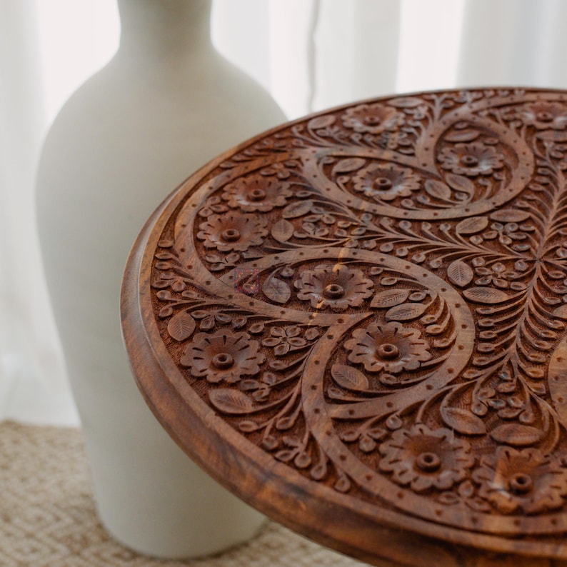 May include: A round, wooden table with intricate floral carvings. The table is a warm brown color and features detailed patterns of flowers and leaves. A white vase is in the background.