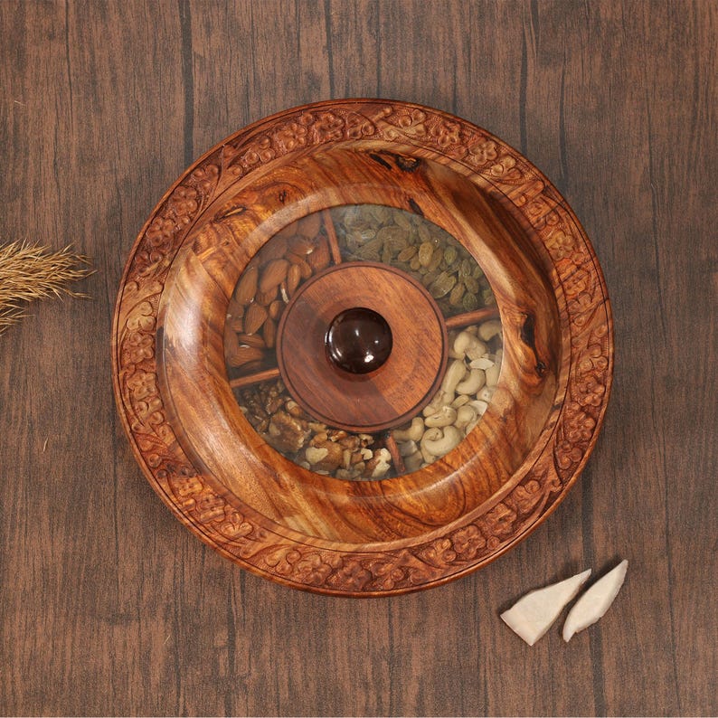May include: A circular wooden serving platter with a central compartment and several sections filled with nuts. The platter has a carved floral design around the edge and a dark brown knob in the centre. The platter is made of a light brown wood.
