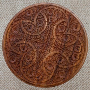 May include: A round wooden table with intricate floral carvings. The table's surface is a rich brown color, with detailed patterns of flowers and leaves carved into the wood. The design is symmetrical and ornate, creating a decorative piece.