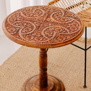 May include: A round, brown wooden side table with intricate floral carvings on the tabletop. The table has a pedestal base and is approximately 24 inches in diameter. The table is made of wood and has a natural finish.