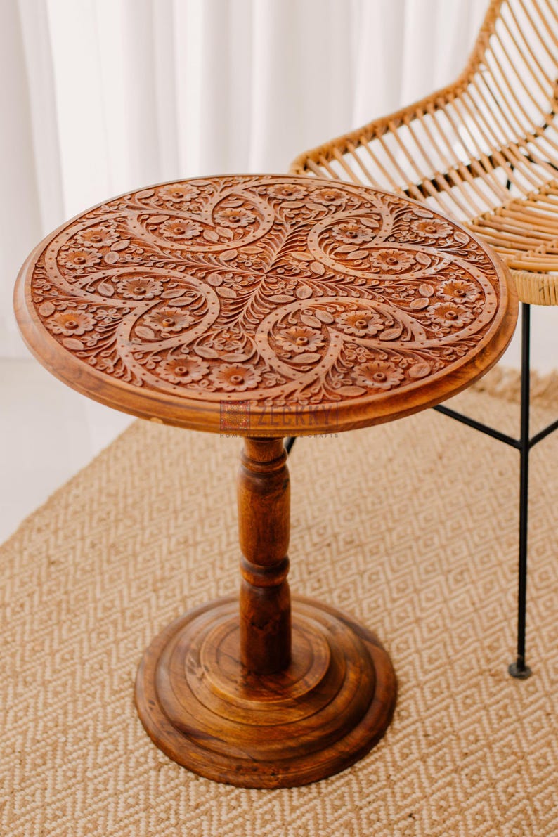 May include: A round, brown wooden side table with intricate floral carvings on the tabletop. The table has a pedestal base and is approximately 24 inches in diameter. The table is made of wood and has a natural finish.
