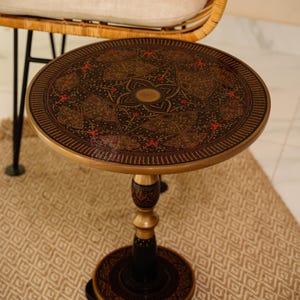 Pu&ograve; includere: Un tavolino rotondo con un design ornato in nero, oro e rosso. Il tavolo ha una base a piedistallo ed &egrave; posto su un tappeto fantasia. Il design del tavolo &egrave; intricato e dettagliato, con una combinazione di colori nero e oro.