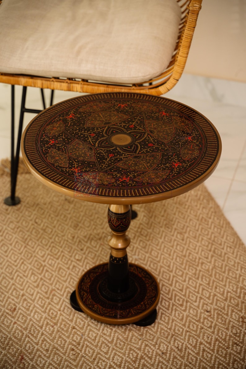 Pu&ograve; includere: Un tavolino rotondo con un design ornato in nero, oro e rosso. Il tavolo ha una base a piedistallo ed &egrave; posto su un tappeto fantasia. Il design del tavolo &egrave; intricato e dettagliato, con una combinazione di colori nero e oro.