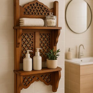 May include: A decorative wooden bathroom shelf unit with a carved geometric pattern. It features a rolled white towel, a glass jar with cotton buds, two white pump bottles, and a small potted plant. The shelf is mounted on a tiled wall.