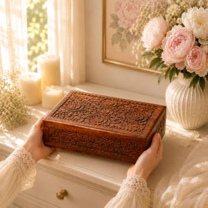 May include: A rectangular wooden box featuring intricate floral carvings across the lid and sides. The dark brown wood displays a detailed relief pattern of flowers and leaves. The box is held by two hands, showcasing its textured surface and craftsmanship.