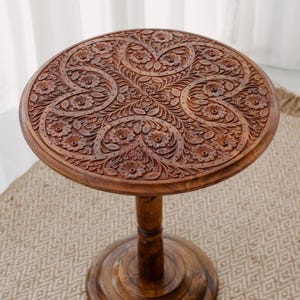 May include: A round, wooden side table with intricate floral carvings on the tabletop. The table is stained a warm brown color and has a pedestal base. The table is approximately 20 inches in diameter and 24 inches tall.