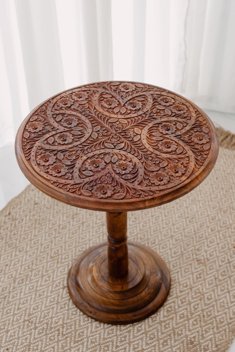 May include: A round, wooden side table with intricate floral carvings on the tabletop. The table is stained a warm brown color and has a pedestal base. The table is approximately 20 inches in diameter and 24 inches tall.