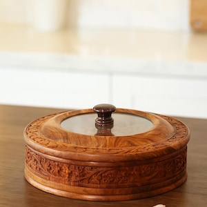 May include: A circular wooden container with a glass lid and a dark brown knob. The container features detailed carved floral patterns. It rests on a wooden surface. The container's diameter is approximately 20 cm.