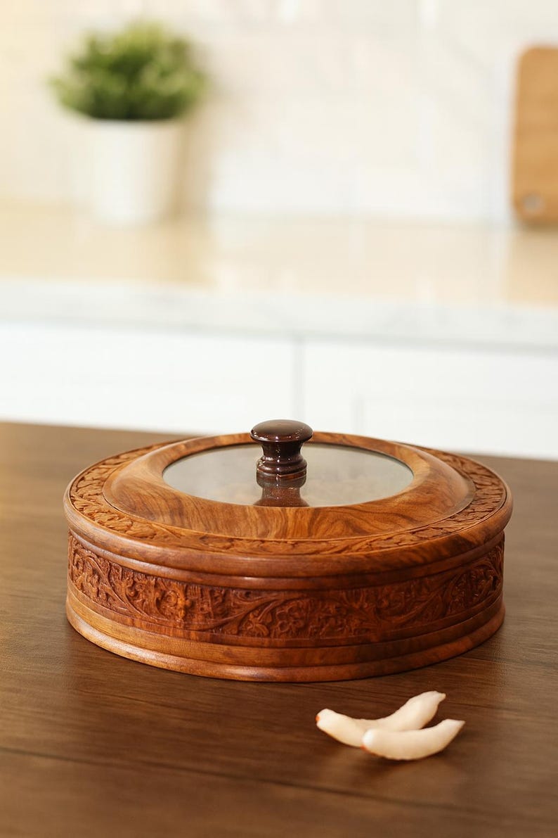 May include: A circular wooden container with a glass lid and a dark brown knob. The container features detailed carved floral patterns. It rests on a wooden surface. The container's diameter is approximately 20 cm.