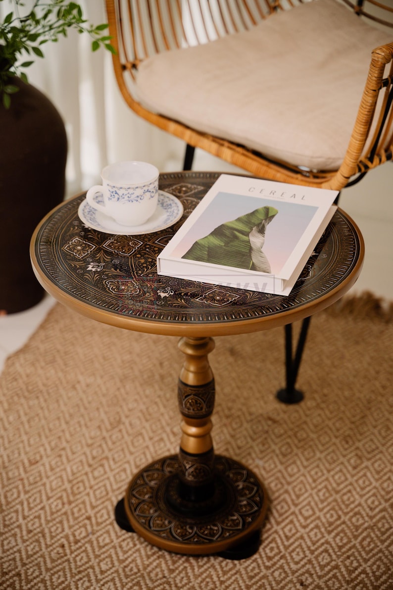Pu&ograve; includere: Un tavolino rotondo e ornato con un bordo color oro e intricati motivi marrone scuro. Una tazza e un piattino bianchi con disegni floreali blu sono sul tavolo, insieme a un libro intitolato "CEREAL". Il tavolo ha un diametro di circa 61 cm.