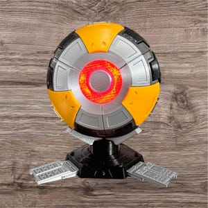 Può includere: Una sfera gialla e argentata con una luce rossa al centro, montata su una base nera con tre piattaforme di atterraggio argentate.