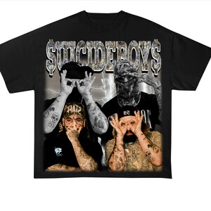 May include: Black t-shirt featuring a graphic of four people with tattoos, making hand gestures. The text "SUICIDEBOYS" is printed across the top in a dollar bill style font. The background has a lightning bolt design.