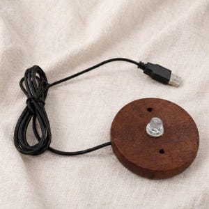 May include: A circular, dark brown wooden base with a clear LED light and a black USB cable. The cable is connected to the base and has a USB connector. The base has two small holes.