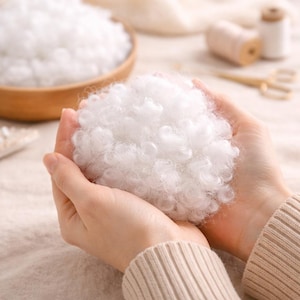 May include: Close-up of fluffy white polyester fiberfill held in cupped hands. The soft, cloud-like material is used for stuffing pillows, cushions, and other craft projects. A wooden bowl filled with more fiberfill is in the background.