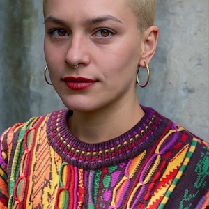 Frau trägt bunten, Vintage gestrickten Pullover im Coogi-Stil mit auffälligem 90er-Jahre-Muster – oversized und unisex, kräftige Farben wie Pink, Gelb, Orange, Lila und Grün. Perfekt für Retro-Liebhaber*innen, Boho- und Streetwear-Fans oder als farbenfroher Festival-Look