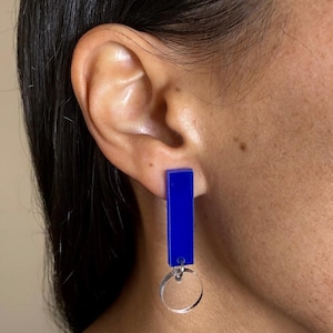Könnte beinhalten: Ein Paar moderne Ohrringe. Jeder Ohrring besteht aus einem leuchtend blauen, rechteckigen Stab, der an einem transparenten, kreisförmigen Anhänger hängt. Die Ohrringe sind für durchbohrte Ohren konzipiert.