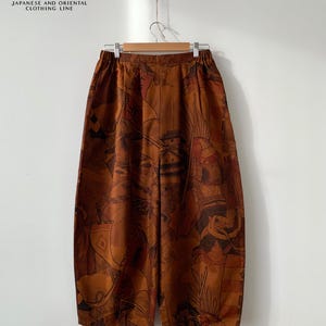 Pode incluir: Calças largas castanhas com uma impressão gráfica castanha escura de guerreiros e arquitetura japonesa tradicional. As calças têm uma cintura elástica e estão expostas num cabide de madeira. O texto SUZU JAPANESE AND ORIENTAL CLOTHING LINE é visível no topo.