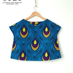 May include: A short-sleeved top featuring a vibrant blue, yellow, and red peacock feather-inspired pattern. The garment is displayed on a wooden hanger. The text SUZU JAPANESE AND ORIENTAL CLOTHING LINE is printed in black in the upper left corner.