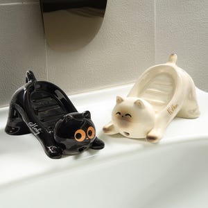 May include: Two ceramic cat-shaped soap dishes. One is glossy black with orange eyes and the name "Marky" on the side. The other is off-white with brown accents and the name "Kitten". Both have a dish for soap.