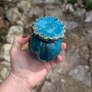 Puede incluir: Un pequeño tarro de cerámica turquesa con tapa a juego. La tapa tiene forma de flor con ribete dorado. El tarro se sostiene en una mano sobre un fondo borroso al aire libre. El tarro mide aproximadamente 7.6 cm de alto.