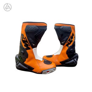 Puede incluir: Par de botas de moto naranjas y negras. Las botas tienen un diseño de caña alta con características de protección, incluyendo áreas reforzadas en los tobillos y espinillas. Las botas presentan el logotipo de KTM y están diseñadas para montar en moto.