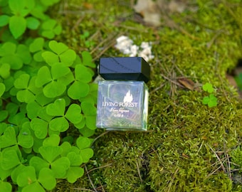 May include: A clear, square glass bottle with a black cap, labelled "Living Forest Home fragrance." The bottle rests on green moss, surrounded by bright green clover. The image evokes a natural, earthy theme.