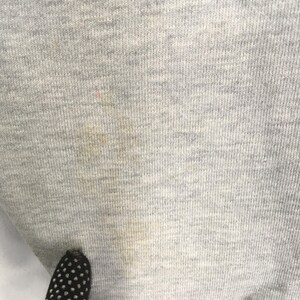 May include: Close-up of a light gray fabric with a ribbed texture. The fabric has a few small stains and a dark object with white polka dots is visible at the bottom. The fabric appears to be a knit material.
